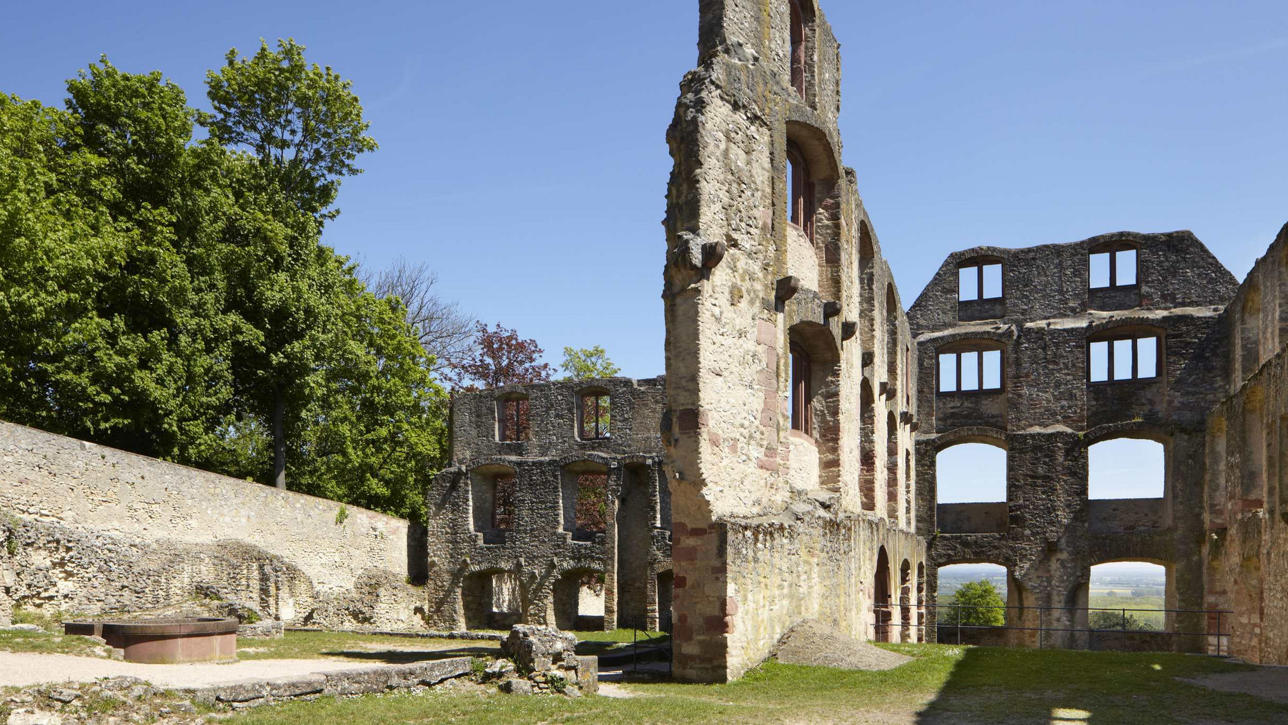 Mauerwände mit Fensteröffnungen, links vorne ein Brunnen, im Hintergrund Bäume